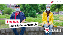 Ein älterer Mann und eine junge Frau sitzen mit Mundschutz auf einer Mauer und unterhalten sich (Verlinkung zu www.niedersachsen.de und Niedersachsen gegen Corona)