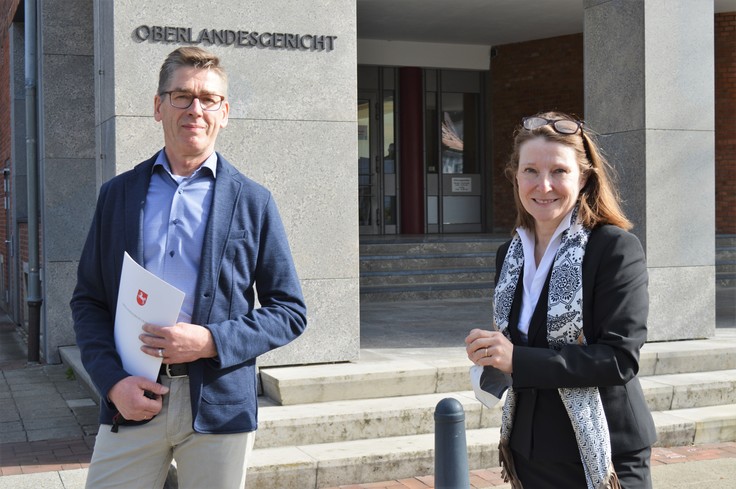 Herr Gehrels und Frau van Hove stehen vor dem Oberlandesgericht. Herr Gehrels hält seine Urkunde zum 50-jährigen Dienstjubiläum in der Hand.